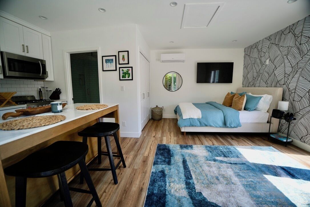 A modern kitchen with white cabinets, a microwave, and a wooden countertop. There is a dining area with stools and a blue rug. A bed with a blue and white blanket is visible in the background.