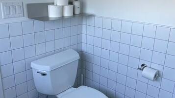 A bathroom with a toilet, a trash bin, and a shelf with toilet paper and a hand soap dispenser.