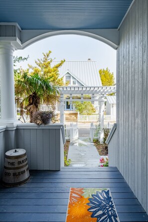 A porch with a wooden barrel, a colorful rug, and a view of a house through a white fence. - Cozy Marshfront Home, walking distance to shops and dining (Bald Head Island)