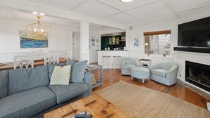 Amplia sala de estar con un sofá gris, una mesa de centro y un televisor. Se aprecia una cocina al fondo con armarios verdes y un comedor con sillas blancas.