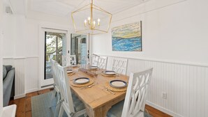 Un comedor con una mesa de madera dispuesta para cuatro personas, sillas blancas, una lámpara de techo geométrica y un cuadro de temática costera en la pared.