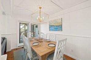 A dining room with a wooden table set for four, white chairs, a geometric chandelier, and a coastal painting on the wall.
