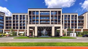 A multi-story hotel with a modern architectural design, featuring a central entrance with a decorative sun-like structure, surrounded by a well-maintained lawn and a parking area. - Wanda Vista Changchun International Movie Metropolis (Changchun)