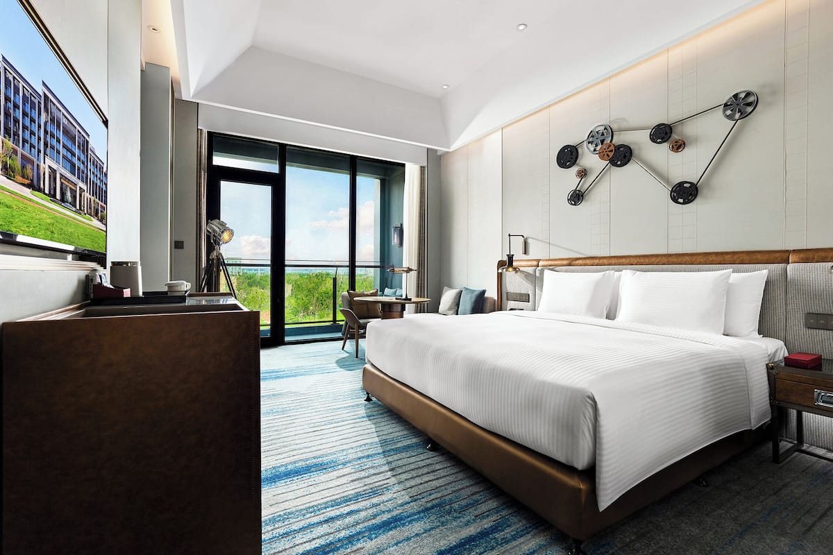A modern hotel room with a large bed, a desk, a chair, and a view of the outdoors.