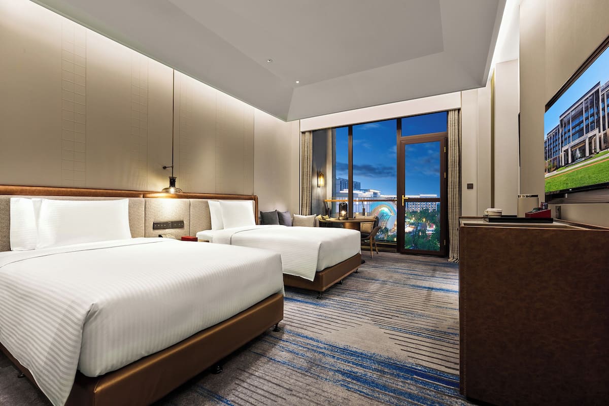 A hotel room with two beds, a desk, a chair, a TV, and a large window with a city view.