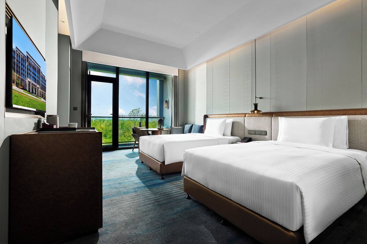 A hotel room with two beds, a desk, a chair, a large window, and a view of greenery.
