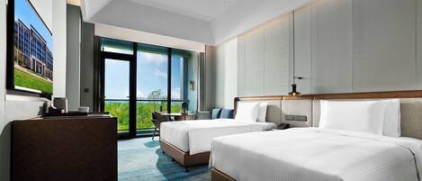 A hotel room with two beds, a desk, a chair, a large window, and a view of greenery.