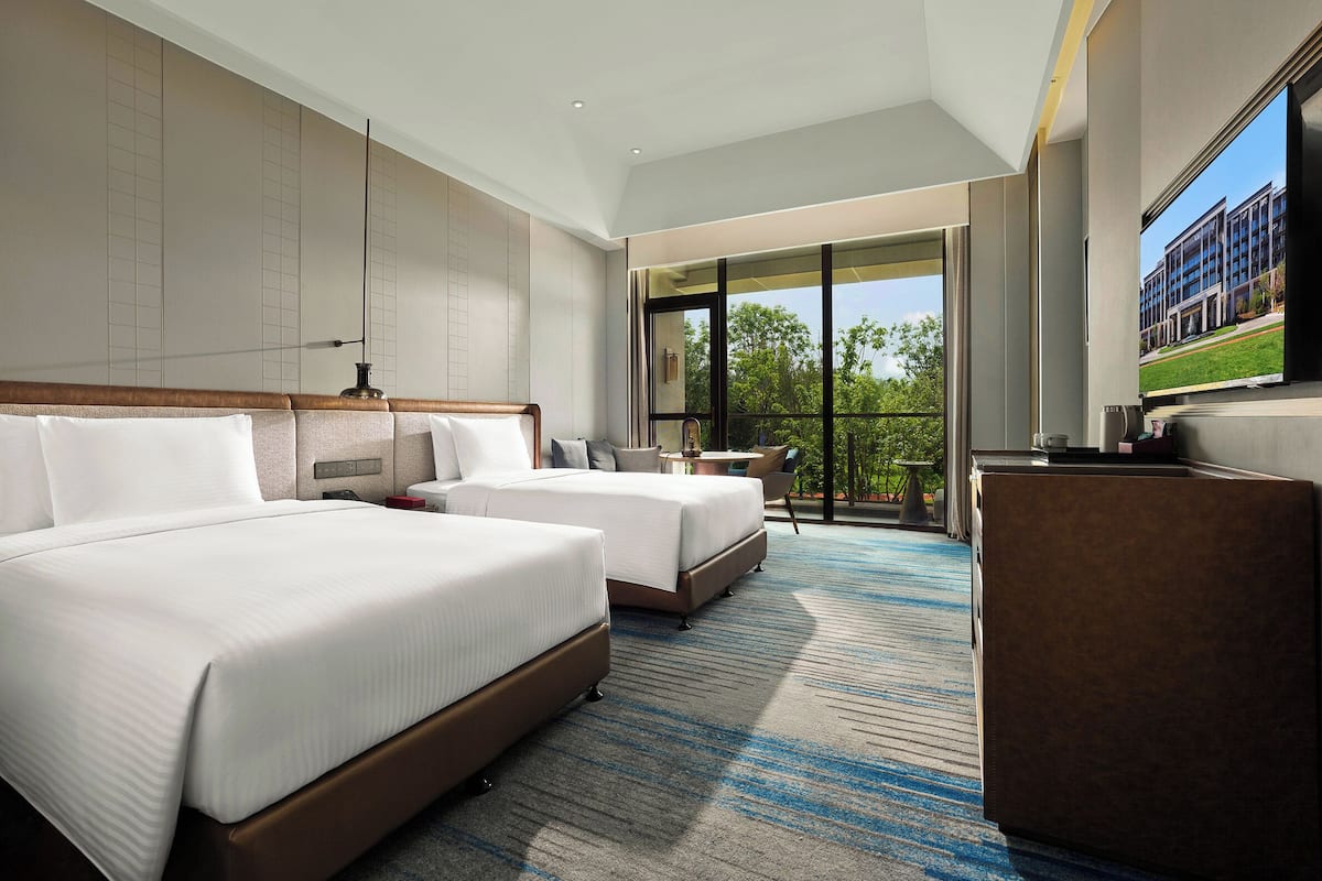 A hotel room with two beds, a desk, a chair, and a large window with a view of buildings and trees.