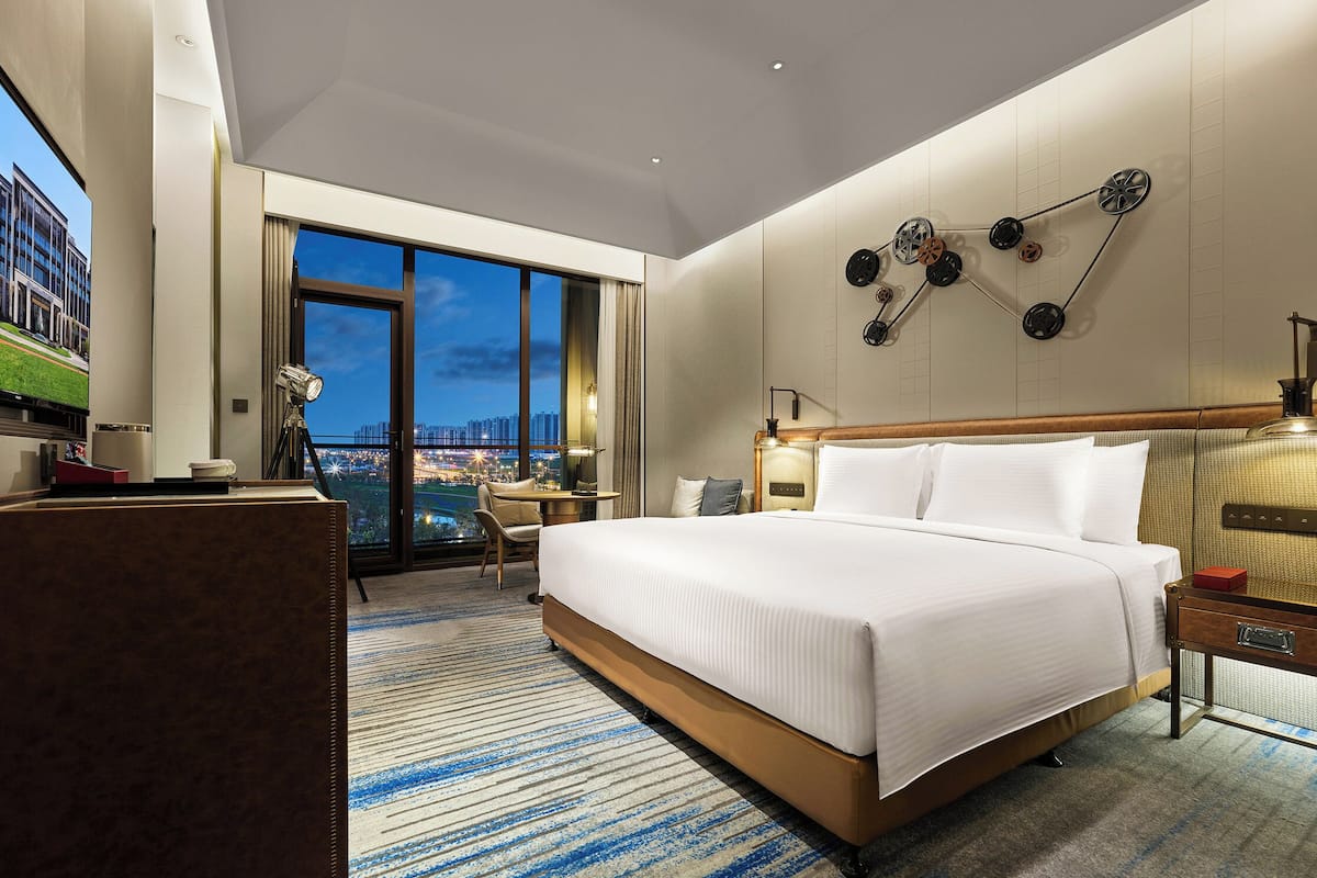 A modern hotel room with a large bed, a desk, a chair, a desk lamp, and a view of the cityscape.