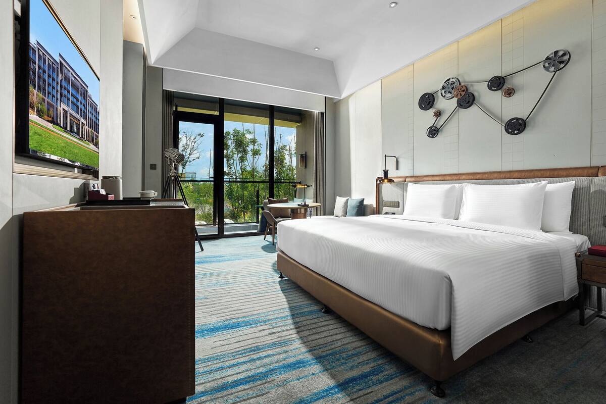 A modern hotel room with a large bed, a desk, and a balcony with a view of the city.