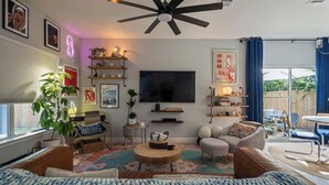 A modern living room with a ceiling fan, a flat-screen TV, and a large window with curtains. - Bohemian Bolthole: Live like a Native Austinite (Austin)