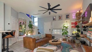 A modern living room with a sofa, coffee table, and bar stools. There are plants, a ceiling fan, and a neon sign. - Bohemian Bolthole: Live like a Native Austinite (Austin)
