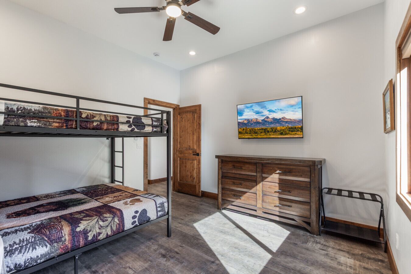 Un lit superposé avec une couvre-lit à motifs, une commode en bois, un ventilateur de plafond et un tableau représentant un paysage, encadré, accroché au mur.