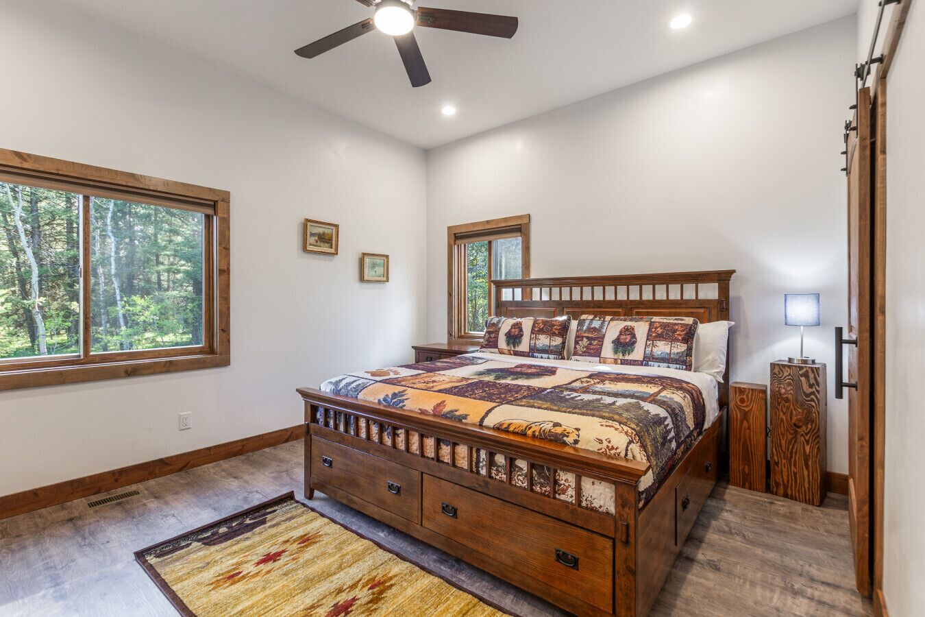 A bedroom with a bed, a ceiling fan, a nightstand, a lamp, and a window with a view of trees.