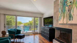 A modern living room with a fireplace, teal furniture, and a large window overlooking a balcony. - Aqua Vie - 4 BR Retreat w/Private Pool, Hot Tub, Elevator (Southern Shores)