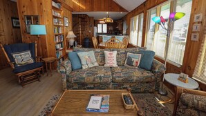 A cozy cabin interior with a patterned sofa, wooden bookshelves, a dining area, and a kitchen. - MacAllister - Semi-Oceanfront 3 BR Beach Bungalow - Dogs Allowed (Southern Shores)