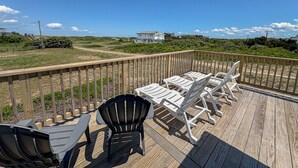 A wooden deck with outdoor furniture, including chairs and tables, overlooking a grassy area with buildings in the distance. - MacAllister - Semi-Oceanfront 3 BR Beach Bungalow - Dogs Allowed (Southern Shores)