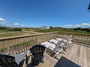 Eine Holzterrasse mit Gartenmöbeln, darunter Stühle und Tische, mit Blick auf eine Rasenfläche und Gebäude in der Ferne.