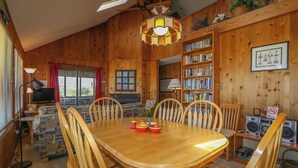 A wooden cabin interior with a dining table, chairs, a bookshelf, and a television. - MacAllister - Semi-Oceanfront 3 BR Beach Bungalow - Dogs Allowed (Southern Shores)