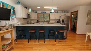Uma cozinha com armários brancos, uma ilha azul com banquetas, eletrodomésticos de aço inoxidável e um letreiro escrito 'Relax'.