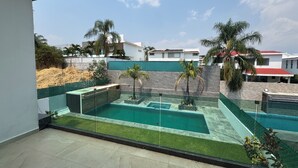 A modern outdoor pool area with a glass barrier, surrounded by a green wall and palm trees. - VACATION FAMILY HOME, MODERN HIMALAYA II (Fraccionamiento Lomas de Cocoyoc)