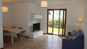 Ein Wohnzimmer mit Fernseher, einer Couch, einem Esstisch mit Stühlen und einer Schiebetür zum Außenbereich.