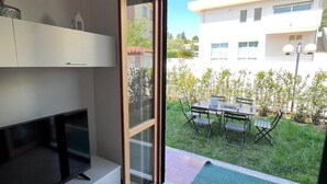 Ein modernes Wohnzimmer mit einem Flachbildfernseher, einer Schiebetür zu einem Balkon mit Essbereich und einem Gebäude im Hintergrund.