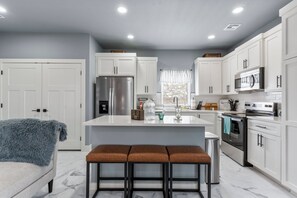 A modern kitchen with white cabinets, a stainless steel refrigerator, a microwave, and an oven. There is a large island with stools, and a window with blinds. - Modern Escape in Downtown Tulsa: Sleeps 12 (Tulsa)