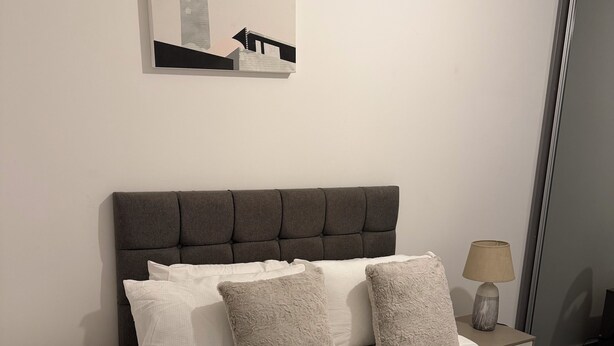 A neatly made bed with a grey upholstered headboard, white linens, and grey throw pillows. A small bedside table with a lamp and a folded white towel is visible. - Stylish Central Southampton 2-Bed Apartment Perfect for all types of Travelers & (Southampton)