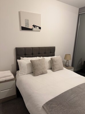 Een net opgemaakt bed met een grijs gestoffeerd hoofdbord, witte lakens en grijze sierkussens. Een klein nachtkastje met een lamp en een opgevouwen witte handdoek is zichtbaar.