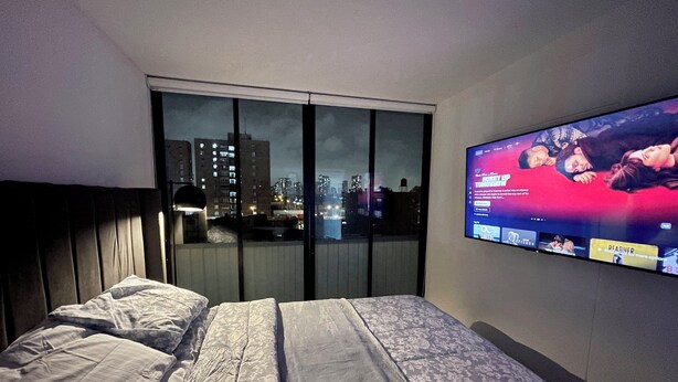 A hotel room with a large bed, a TV mounted on the wall, and a view of a cityscape through a large window. - Digital Nomad, Expat and Foreign Student Haven! (Pueblo Libre)