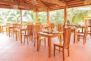 Un restaurante con mesas y sillas de madera, dispuesto en un amplio espacio exterior con suelo de baldosas y techo de paja.