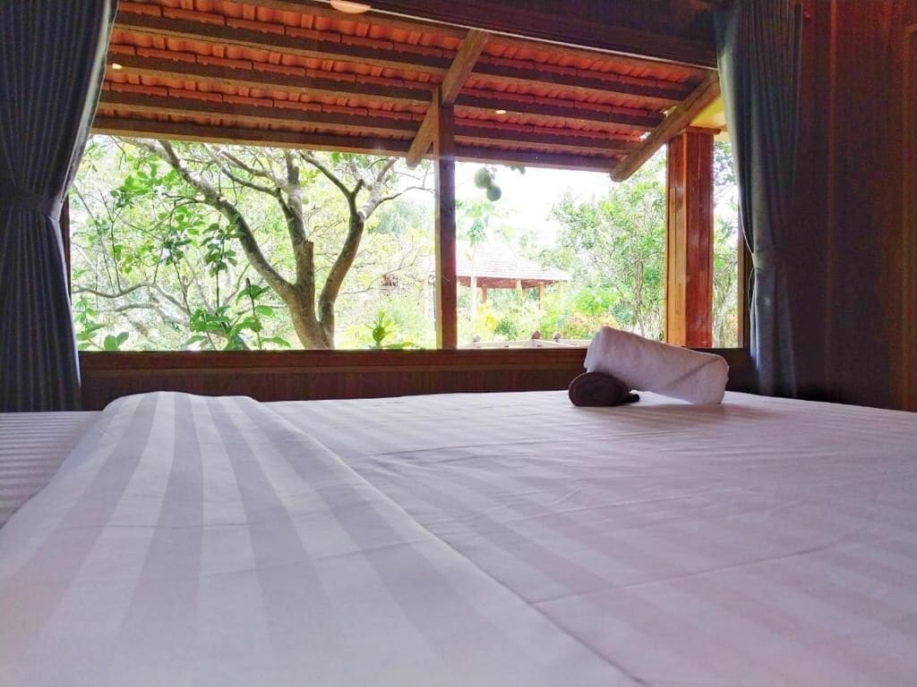 Una cama con sábana blanca y almohada, techo de madera y ventana con vista a árboles y un edificio exterior.