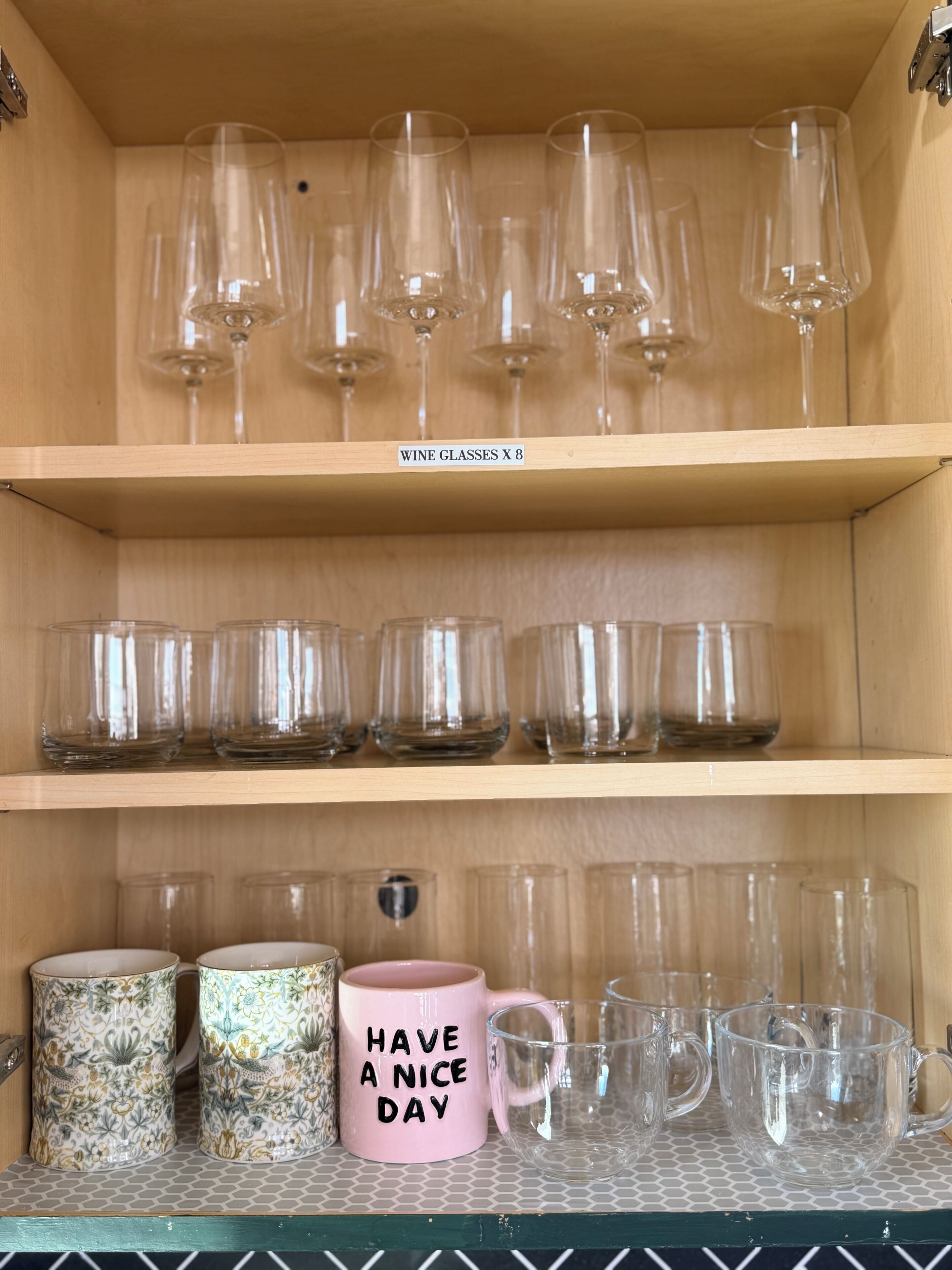 A shelf with wine glasses and clear mugs, and a pink mug with 'HAVE A NICE DAY' text.
