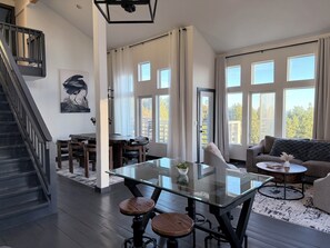 Ein modernes Wohnzimmer mit einem Esstisch aus Glas, einer Treppe und großen Fenstern mit Blick ins Freie.