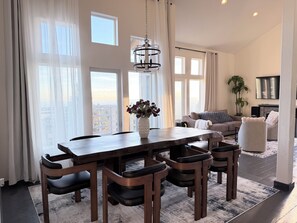 Ein geräumiges Wohnzimmer mit einem großen Esstisch, bequemen Sitzgelegenheiten, einem Kamin und großen Fenstern mit Blick auf die Stadt.