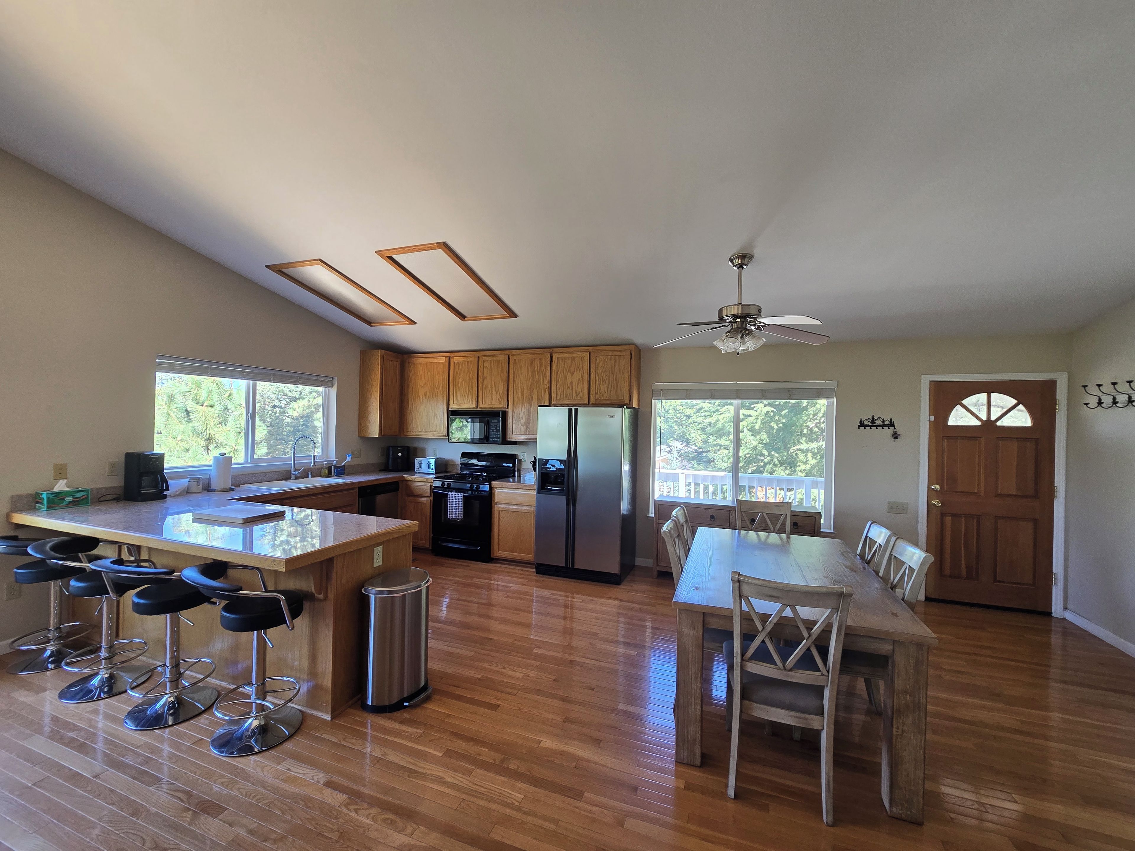 A spacious kitchen with wooden cabinets, a central island with bar stools, a dining table with chairs, and a large window providing a view of the outdoors.