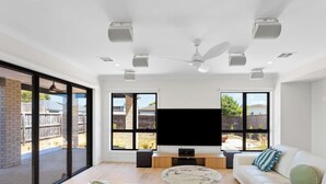 Ein geräumiges Wohnzimmer mit großen Fenstern, einem Deckenventilator und einem Flachbildfernseher.