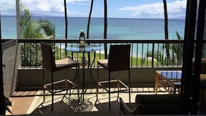 A balcony with a table and chairs, overlooking a beach with palm trees and the ocean. - OCEANFRONT with Private Beach! Hale Mahina A206 (Lahaina)