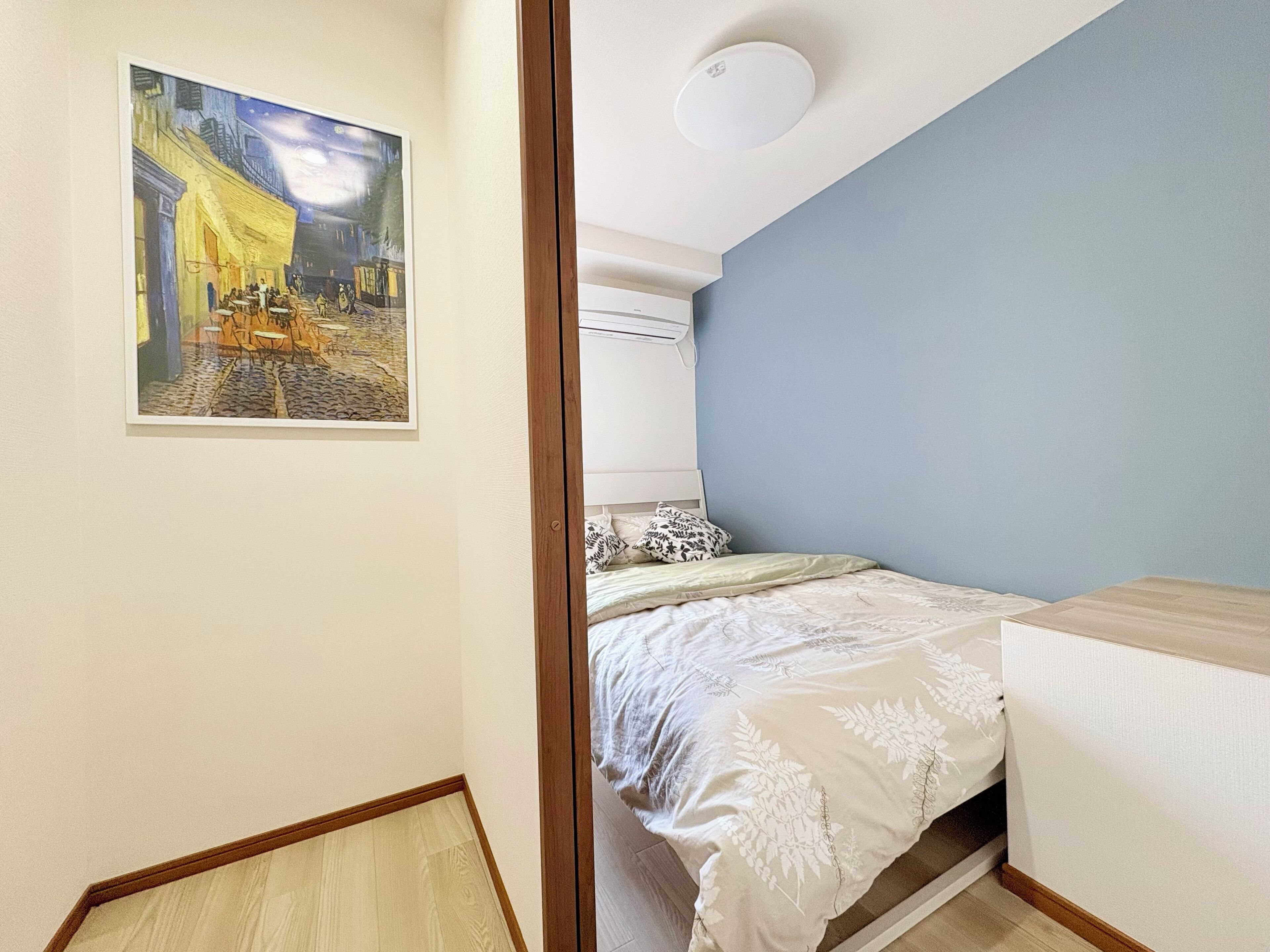 A bedroom with a bed, a nightstand, a wall-mounted air conditioner, and a painting of a café scene.