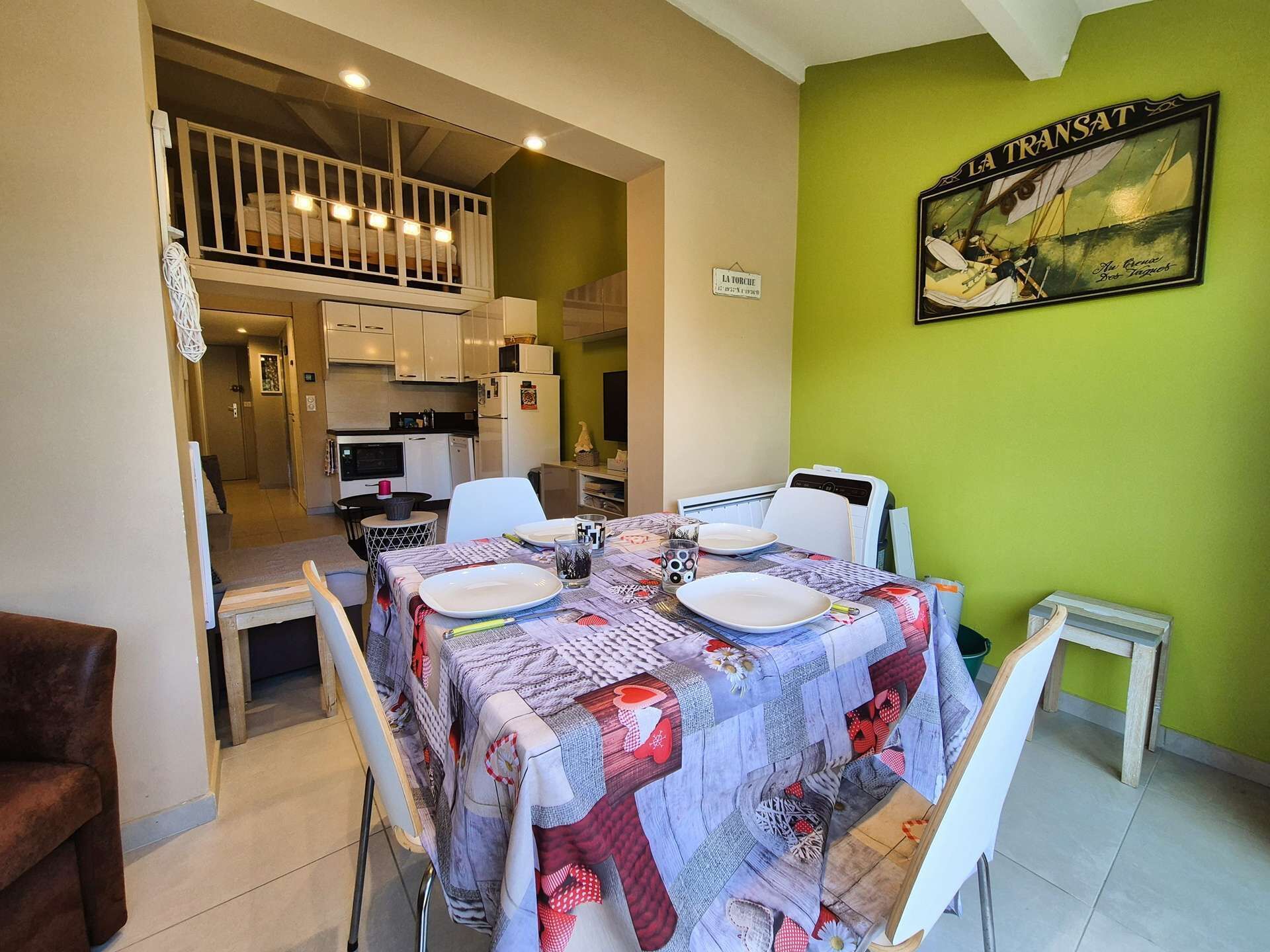 Zona pranzo con tavolo apparecchiato per due, cucina sullo sfondo e parete verde con un cartello.