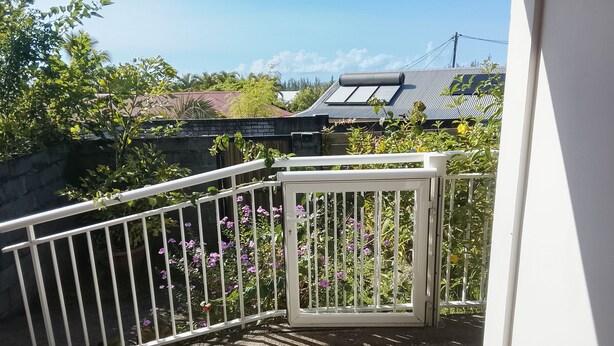 A white metal gate leading to a balcony with a view of a residential area, including houses and trees. - Charming room in St Philippe, between the sea and the mountains, close to the GRR2 footpaths. (Saint-Philippe)