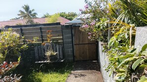 A narrow concrete pathway leading to a wooden gate, surrounded by lush greenery and a wooden fence. - Charming room in St Philippe, between the sea and the mountains, close to the GRR2 footpaths. (Saint-Philippe)