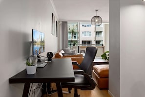 Ein modernes Wohnzimmer mit Schreibtisch, großem Flachbildfernseher, bequemer Couch und Sessel. Auf dem Schreibtisch steht ein Blumentopf und eine Pendelleuchte ist angebracht.