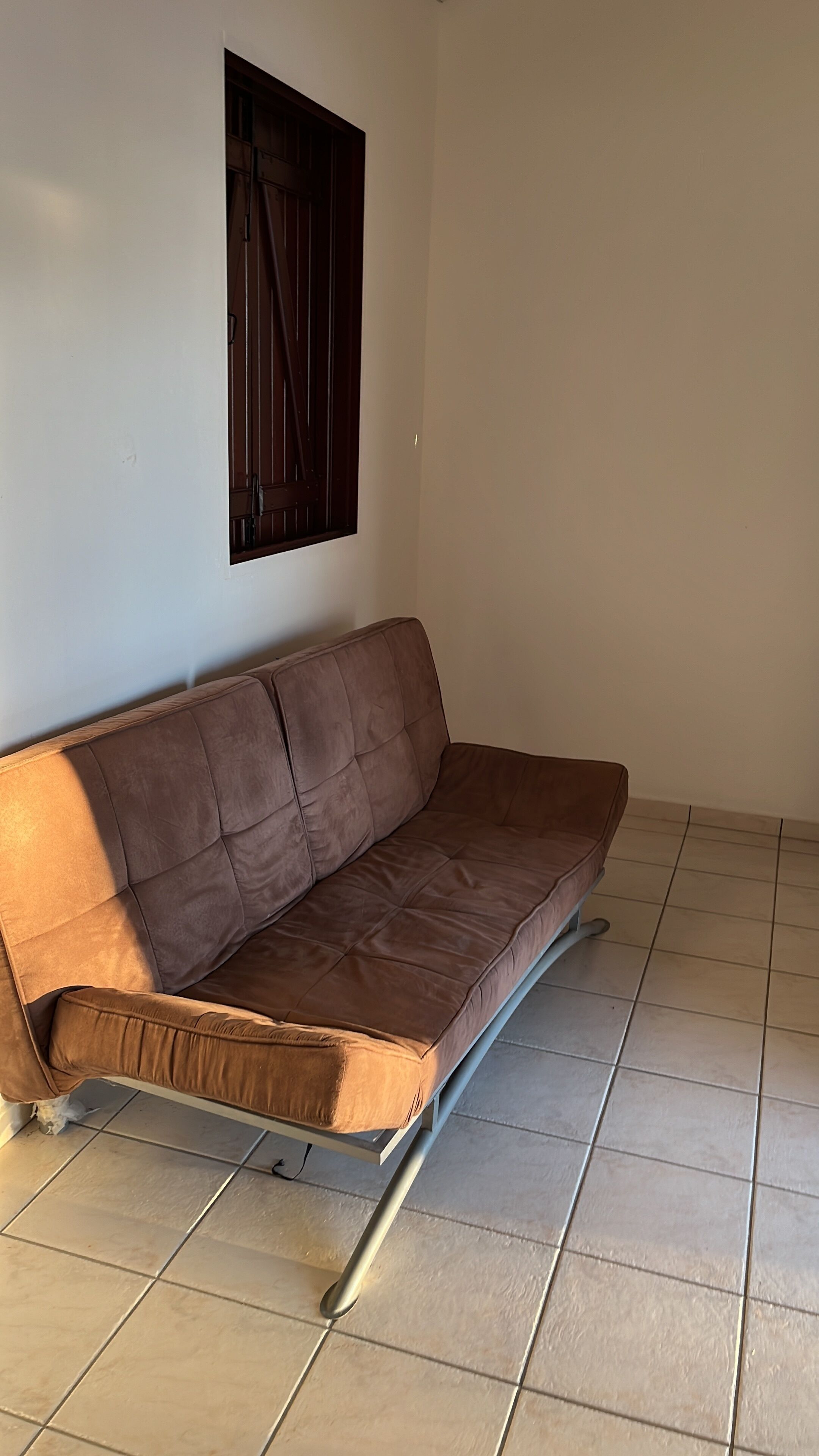 Een bruine bank met metalen poten in een kamer met betegelde vloer en een raam met donkere gordijnen.