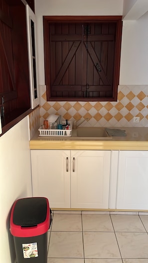 A kitchen with white cabinets, a wooden door, a sink, and a trash bin. - Between sea and sky (Deshaies)