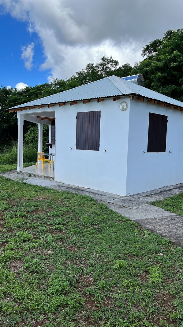 A small, single-story building with a corrugated metal roof, a covered porch with a yellow chair, and a small window.