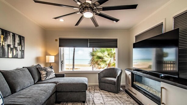 Un salón con sofá modular, una butaca, televisor de pantalla plana, ventilador de techo, ventana grande con vista a la playa y suelo estampado.