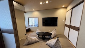 Una habitación con esteras de tatami, una mesa de tatami, dos cojines de tatami, un televisor y una ventana con persianas.
