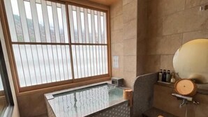 Un baño de estilo japonés tradicional con suelo de madera, una bañera de hidromasaje envuelta en vidrio, una pared de piedra y un taburete de madera.